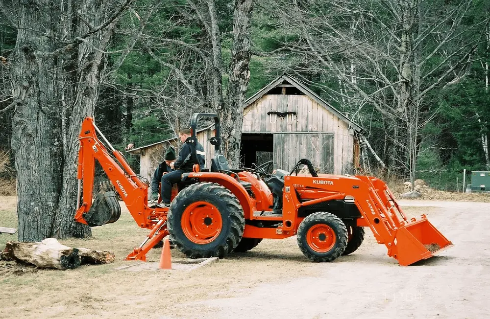 Orange Kubota compact tractor with front loader and rear backhoe parked by a wooden barn